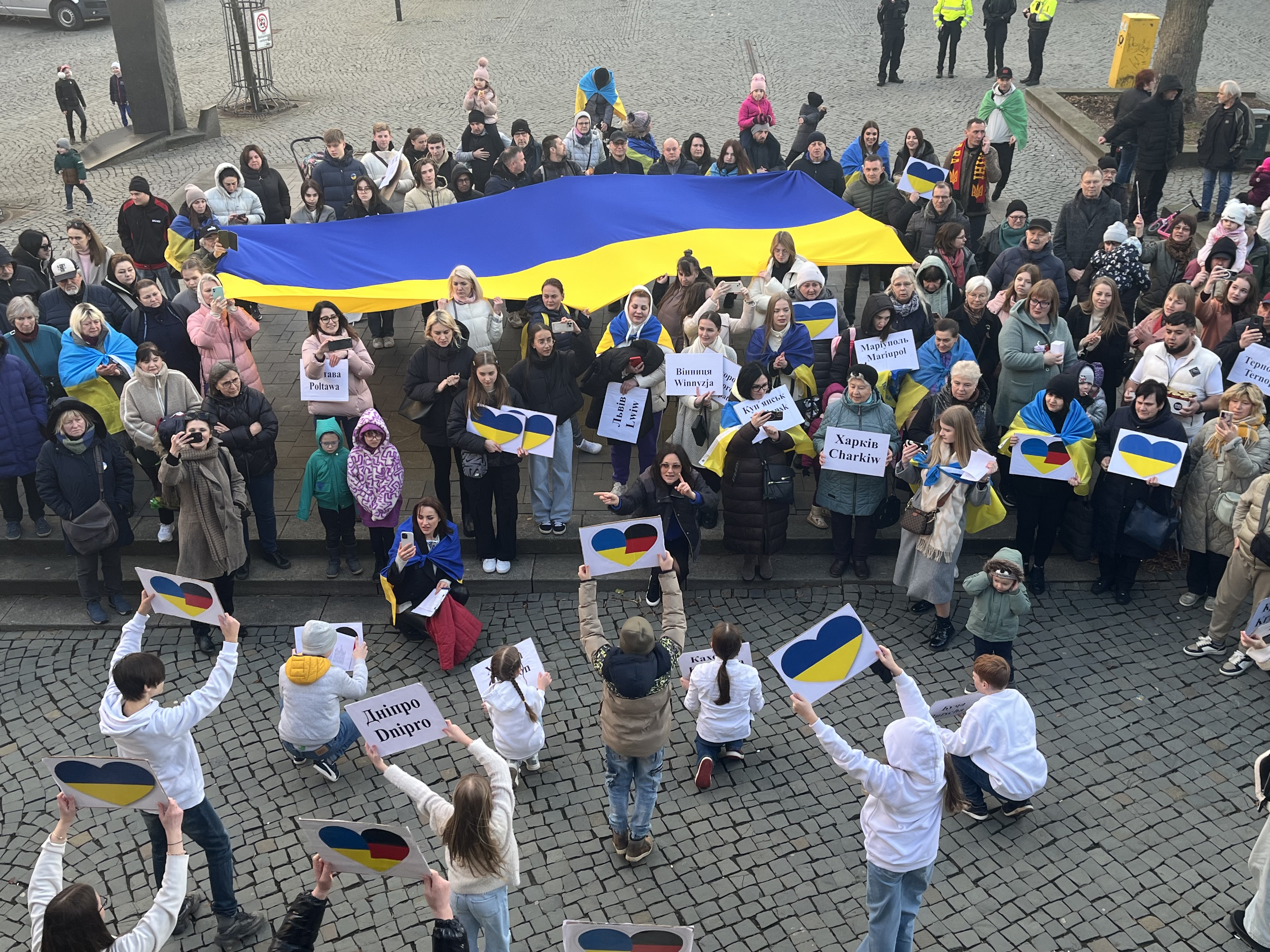 Blick von oben: Blick von der Alten Wache am 24.02.2024, dem Jahrestag des russischen Überfalls. Auf dem Marktplatz stehen viele Menschen mit Schildern und einer großen Ukrainefahne. Die Schilder enthalten entweder Text, oder Herzen in den Farben der Ukraine und manchmal auch zusätzlich den von Deutschland.